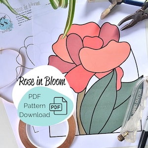 Peut inclure: Un modèle PDF imprimable pour une rose en fleurs en vitrail. Le modèle comprend une rose aux pétales roses et aux feuilles vertes. L'image montre également des outils pour la fabrication de vitraux, notamment un coupe-verre, du ruban adhésif en cuivre et un marqueur noir.