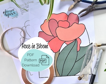 Op de afbeelding: Een printbaar PDF-patroon voor een bloeiende roos van glas-in-lood. Het patroon bevat een roos met roze bloemblaadjes en groene bladeren. De afbeelding toont ook gereedschap voor glas-in-lood, waaronder een glascutter, koperfolietape en een zwarte marker.