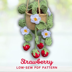 May include: A crocheted hanging planter with a strawberry theme. The beige basket holds green leaves and white flowers with yellow centers. Red strawberries with green tops hang below. The text "Strawberry LOW-SEW PDF PATTERN" is displayed.