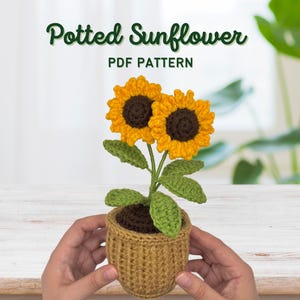Könnte beinhalten: Eine gehäkelte Topfsonnenblume mit zwei gelb-braunen Blüten, grünen Blättern und einem braunen Topf. Der Text "Potted Sunflower PDF Pattern" steht oben. Der Topf wird von zwei Händen gehalten.