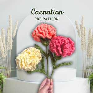 Puede incluir: Un ramo de claveles de ganchillo hechos a mano con tres flores rosas y amarillas, con tallos y hojas verdes. El texto "Carnation PDF Pattern" se muestra en la parte superior. Las flores se exhiben sobre un pedestal blanco.