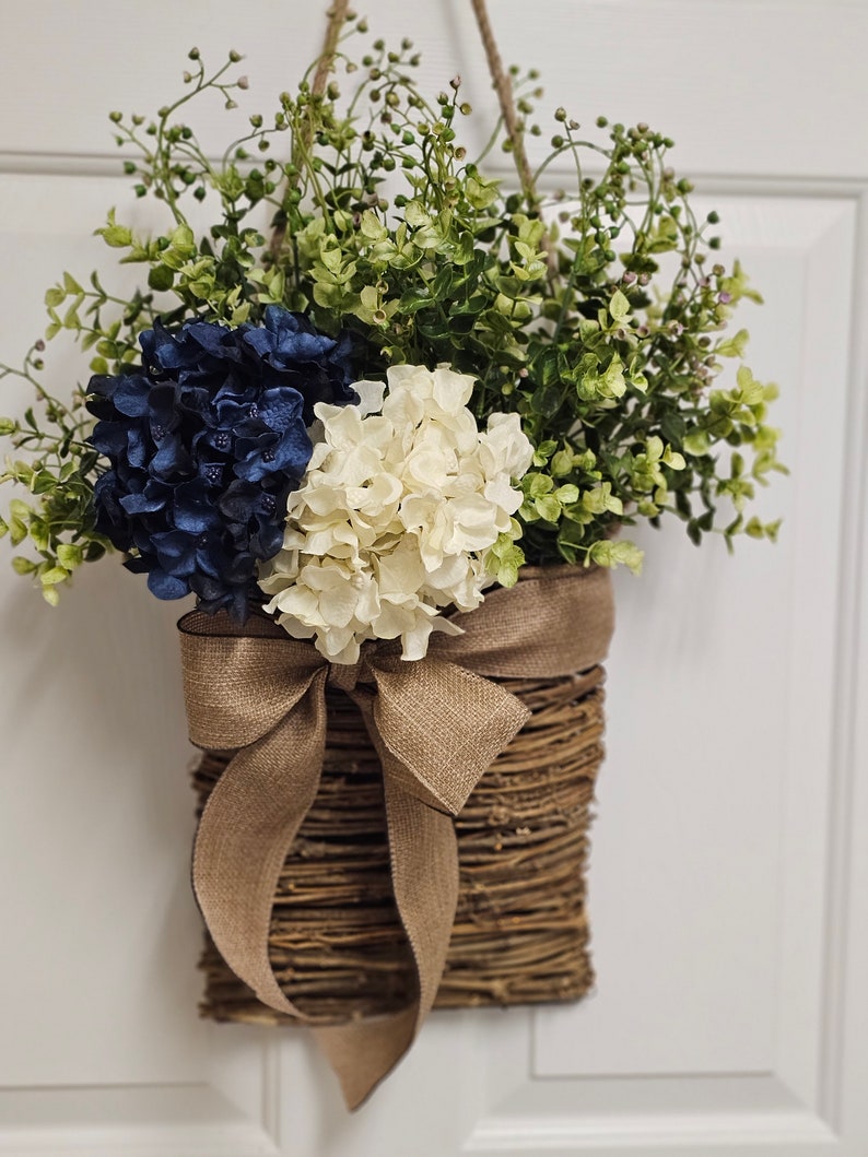 May include: A woven wicker basket with a hessian bow hangs on a white door. The basket is filled with blue and white hydrangeas and green foliage.