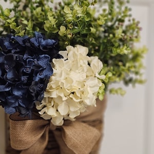 May include: A brown woven basket with a hessian bow, filled with white and navy blue hydrangeas and green foliage.