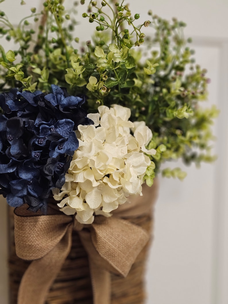 May include: A brown woven basket with a hessian bow, filled with white and navy blue hydrangeas and green foliage.