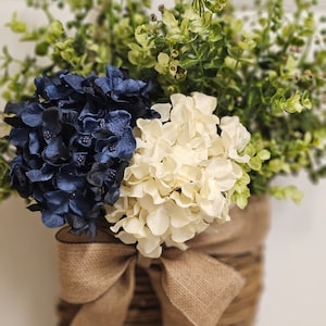 May include: A woven basket with a hessian bow holds a bouquet of blue and white hydrangeas and green foliage.