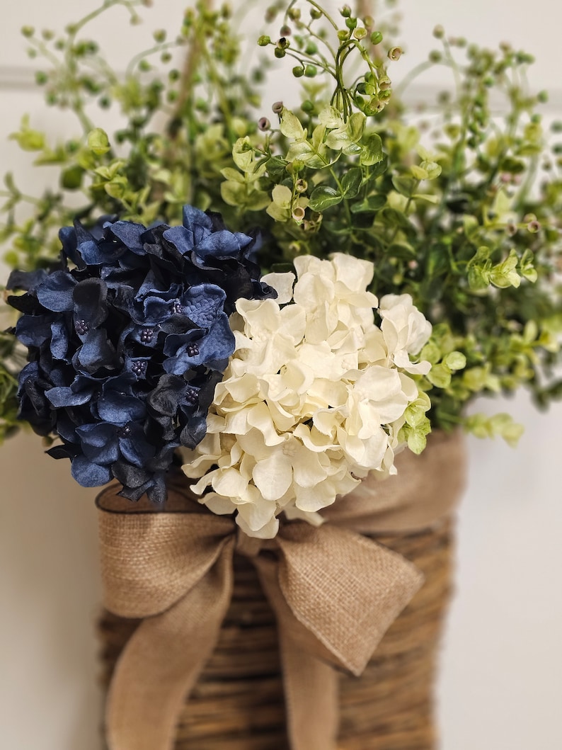 May include: A woven basket with a hessian bow holds a bouquet of blue and white hydrangeas and green foliage.