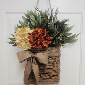 May include: A decorative hanging basket filled with faux flowers and greenery. The basket is made of woven twigs and features a burlap ribbon bow. The flowers include orange, yellow, and green hues, creating a warm, inviting aesthetic.