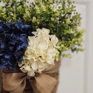 May include: A rustic brown wicker basket filled with blue and white hydrangeas and green foliage, tied with a burlap bow.