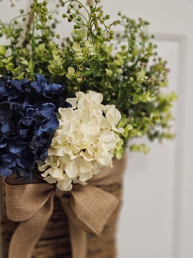 May include: A rustic brown wicker basket filled with blue and white hydrangeas and green foliage, tied with a burlap bow.