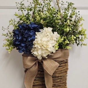 May include: A woven wicker basket with a brown hessian bow hangs from a white door. The basket is filled with blue and white hydrangeas and green foliage.