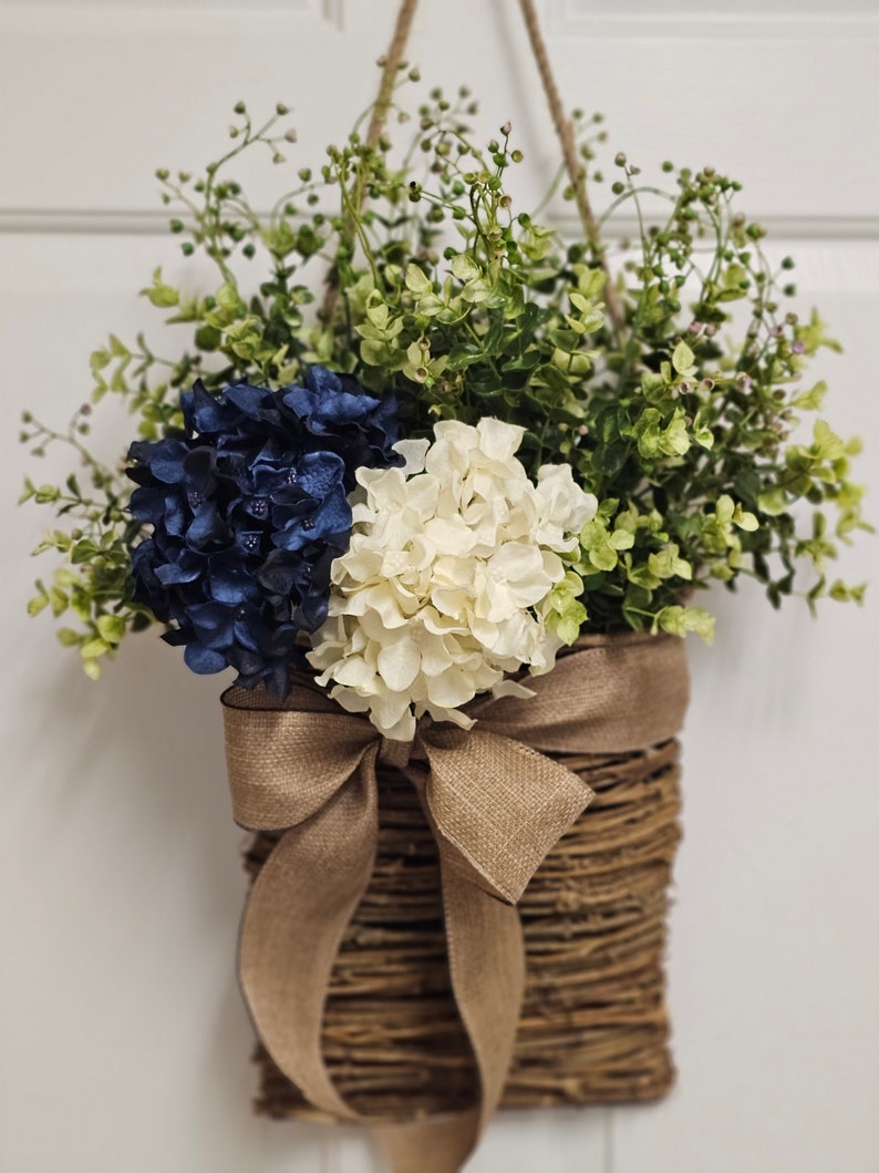May include: A woven wicker basket with a brown hessian bow hangs from a white door. The basket is filled with blue and white hydrangeas and green foliage.