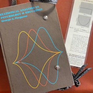 May include: A brown hardcover book with a black leather strap. The book cover features a colorful graphic design of three overlapping waves. The title of the book is "Statistical Analysis in Psychology and Education" by George A. Ferguson.