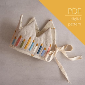 May include: A beige fabric crown with a tie closure, decorated with colorful embroidered candles. PDF digital pattern.