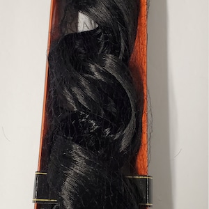 May include: A black hair extension in a box with a gold label. The hair extension is long and wavy. The label says "Gel Throwaway Do Not Eat Desiccant".