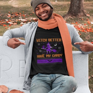 Könnte beinhalten: Ein schwarzes T-Shirt mit einem lila und weißen Design, das eine Hexe auf einem Besen zeigt. Der Text auf dem Shirt lautet "Witch Better Have My Candy".