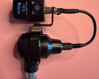 May include: A black and silver tattoo machine with a digital power supply unit attached. The display shows the text AMBITION, 0.0v, and a battery icon. The device features a black control panel with plus, minus, and circular buttons, connected by a black cable to the main body.