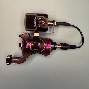 May include: A rotary tattoo machine with a distressed purple and black finish, featuring a black battery pack labeled AMBITION with plus, minus, and circular control buttons. A black power cord connects the battery pack to the machine body.