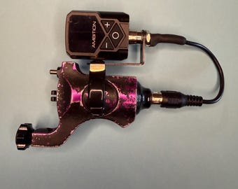 May include: A rotary tattoo machine with a distressed purple and black finish, featuring a black battery pack labeled AMBITION with plus, minus, and circular control buttons. A black power cord connects the battery pack to the machine body.