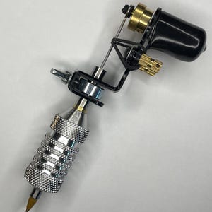 May include: A professional tattoo machine with a black motor housing, gold-colored accents, and a silver, textured grip. The needle tip is visible, and the machine is set against a white background.