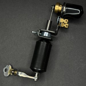 May include: A black and silver tattoo machine with a ribbed grip, brass accents, and a curved black handle. The machine is set against a dark gray background, showcasing its intricate mechanical design.