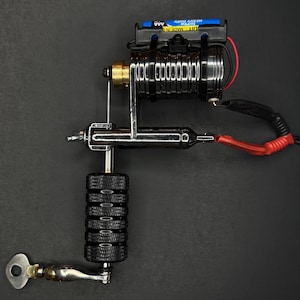 May include: A professional tattoo machine with a black and silver metal frame. The machine has a black grip, a silver needle, and a black power supply with a blue battery. The machine is on a black surface.