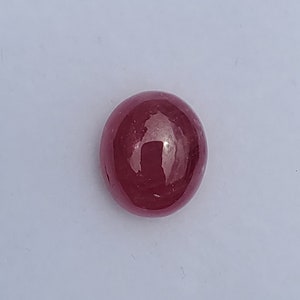 May include: A polished, oval-shaped, deep red gemstone. The gem has a smooth, reflective surface, with a slight sheen. The stone is set against a plain white background.