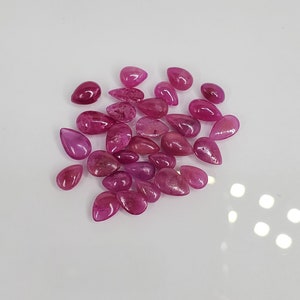 May include: A collection of polished, teardrop-shaped gemstones in varying shades of pink and red. The gems are scattered on a white surface, showcasing their smooth, reflective surfaces and natural variations in color and clarity.