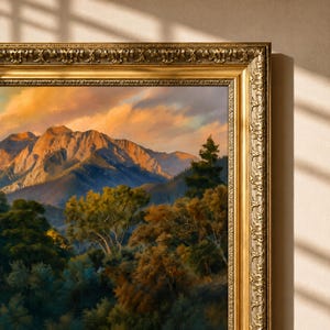 May include: A landscape painting in an ornate, antique-style gold frame. The artwork depicts a mountain range under a sunset sky with orange and yellow clouds, overlooking a dense forest with green and brown trees in the foreground.