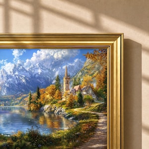 May include: A framed landscape painting featuring a tall church spire nestled among autumn trees, a calm lake, and snow-capped mountains under a bright blue sky. The artwork is encased in a polished gold-toned frame with a classic molding profile.