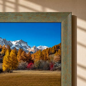 May include: A framed landscape photograph featuring snow-capped mountains under a clear blue sky, with a foreground of golden autumn trees and a dry meadow. The frame is made of wood with a distressed, weathered green and brown textured finish.