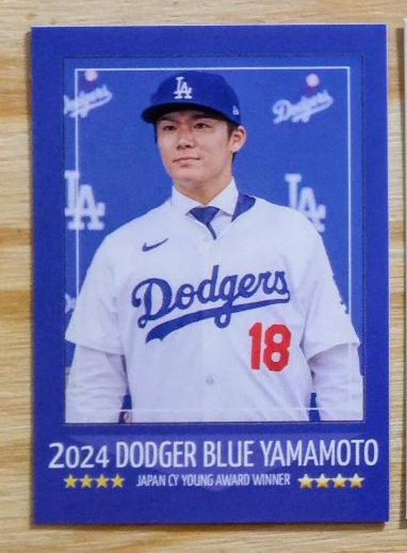 May include: A baseball card featuring a man in a blue Dodgers baseball cap and jersey with the number 18. The card has the text "2024 DODGER BLUE YAMAMOTO" and "JAPAN CY YOUNG AWARD WINNER".