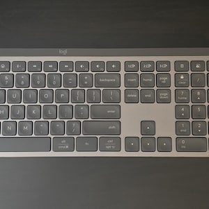 May include: A wireless gray keyboard with a full numeric keypad and a matching gray wireless computer mouse. Both devices feature the Logi brand logo. The keyboard has a standard QWERTY layout with function keys and a metallic finish frame.