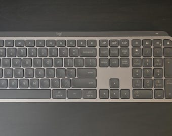 May include: A dark gray wireless keyboard with a full QWERTY layout and a matching dark gray wireless computer mouse. Both devices feature the Logi logo in a light gray color. The keyboard includes a numeric keypad and function keys.