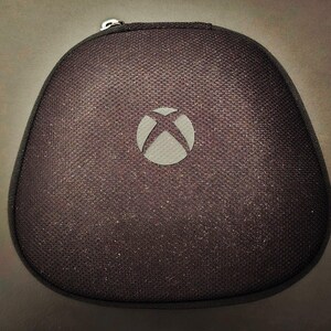 May include: A black, textured Xbox controller case with a zip closure and silver pull. The case is oval-shaped with the Xbox logo in a grey circle on the front. The case is designed to protect the controller.