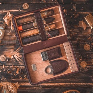 May include: A brown leather cigar travel case containing five cigars of varying shades, a metal cigar cutter, and a lighter. The interior is lined with light-colored wood, featuring dedicated slots for the cigars and accessories, with a dark brown textured leather exterior.