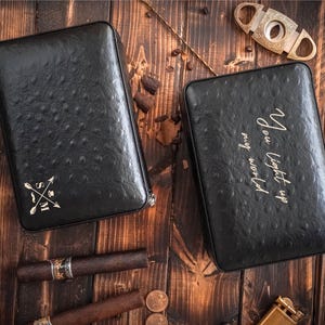 May include: Two black leather cigar cases with textured surfaces are displayed on a wooden table. One case has an arrow and cross design, the other has the text "You light up my world". Cigars, a cigar cutter, and coins are also visible.