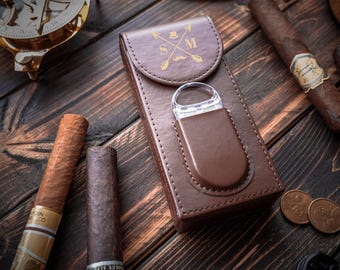 May include: A brown leather cigar case with a gold emblem and a cigar cutter. Several cigars and coins are arranged on a dark wooden surface. A sundial is visible in the background.