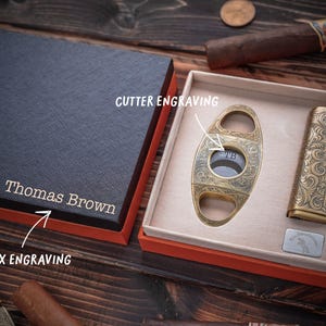 Peut inclure: Un coffret cadeau noir avec le nom Thomas Brown grav&eacute; sur le couvercle, contenant un coupe-cigare dor&eacute; avec la gravure TB et un briquet dor&eacute; avec la gravure Thomas, tous deux orn&eacute;s de motifs &agrave; volutes complexes sur leurs surfaces.