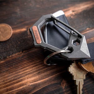 May include: A grey metal multi-tool keyring with a copper-coloured accent, engraved with the word "Hubby". The keyring is attached to a black strap and holds several keys. A coin and a black object are also visible.