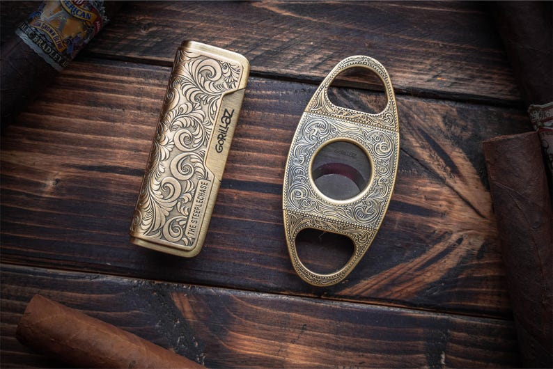 Peut inclure: Un briquet rectangulaire couleur laiton avec des gravures complexes en volutes et le texte GORILLAZ et THE STEEPLECHASE, accompagn&eacute; d'un coupe-cigare ovale couleur laiton avec des motifs floraux assortis et une ouverture circulaire centrale.