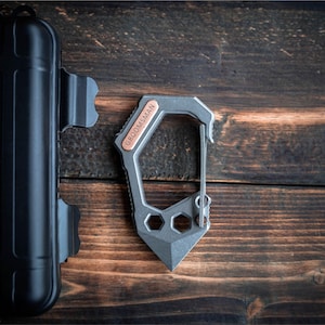 May include: A gray multi-tool carabiner with a copper accent that reads "GROOMSMAN". The tool has a built-in knife and hexagonal cutouts. A black case and a gold coin are also visible.