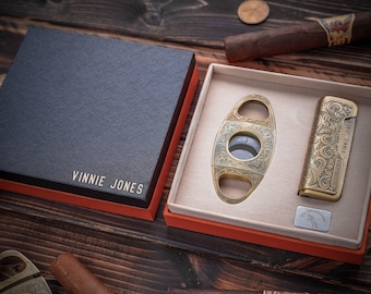 May include: A luxury cigar set featuring a navy blue box with the text "VINNIE JONES" in gold lettering. Inside, a gold cigar cutter and lighter with ornate designs are displayed. The set is presented on a wooden surface.
