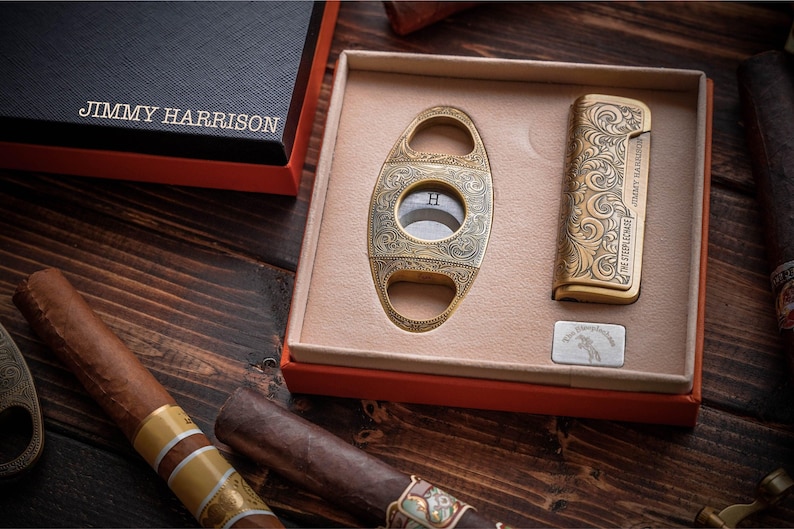 May include: A luxury cigar set featuring a gold-toned cigar cutter and lighter with ornate detailing, presented in a red-trimmed box. The set includes a silver metal plate and is accompanied by several cigars on a wooden surface.