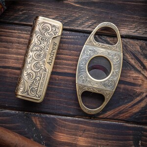 Peut inclure: Un briquet rectangulaire en laiton avec un motif complexe de volutes, le nom Andrew grav&eacute; et le texte The Steeplechase, accompagn&eacute; d'un coupe-cigare ovale en laiton avec des volutes assorties et une ouverture circulaire centrale.