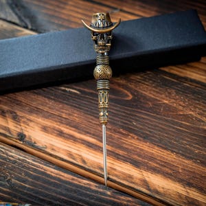 May include: A decorative letter opener with a skull-shaped handle wearing a cowboy hat. The handle is bronze-colored with intricate detailing. The blade is silver and the opener rests on a wooden surface with a black box in the background.