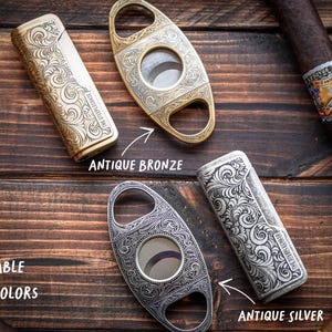 Peut inclure: Deux briquets rectangulaires et deux coupe-cigares ovales avec des gravures &agrave; volutes complexes. Un ensemble est fini en bronze antique et l'autre en argent antique. Les &eacute;tiquettes indiquent : Available in two colors, Antique Bronze, Antique Silver.