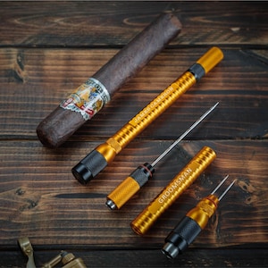 May include: An overhead shot of a cigar and several gold-colored cigar tools on a dark wooden surface. The tools include a cigar awl and a cigar poker, with the text "JAMES CAMERON" and "GROOMSMAN" engraved on them. A cigar cutter is visible.