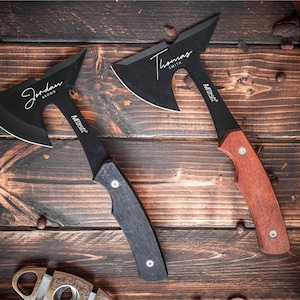 May include: Two black hatchets with personalized names, "Jordan Brown" and "Thomas Smith", are displayed on a wooden surface. One hatchet has a black handle, and the other has a reddish-brown handle. The hatchets have a sharp, angled blade.