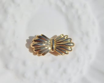 Puede incluir: Un broche de color dorado en forma de lazo con un diseño texturizado.