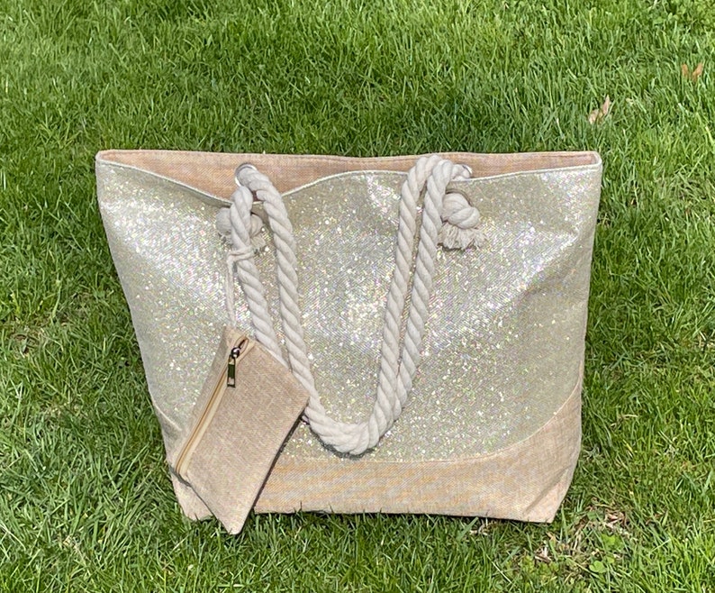 May include: A large, white glitter tote bag with a beige canvas bottom and rope handles. The bag has a smaller beige canvas pouch attached to the side.